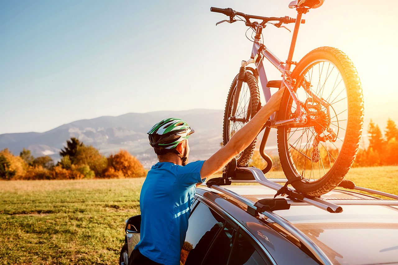 Man in blue T-shirt putting mountain bike on car roof rack. Low sun over background hills.